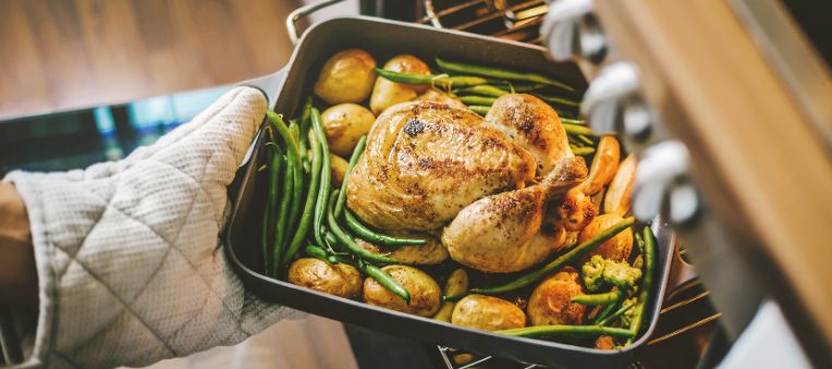 A person with oven mitts is pulling a pan with cooked chicken, potatoes, and beans out of the oven.